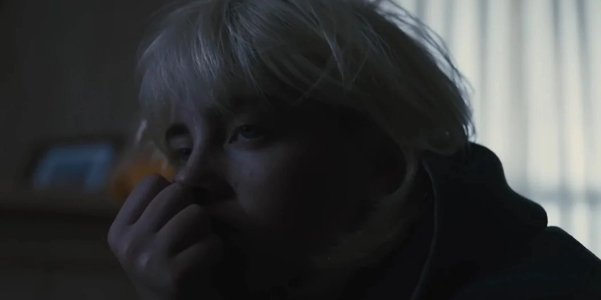 Billie Eilish sitting alone in dim lighting with a distant and emotionally exhausted expression