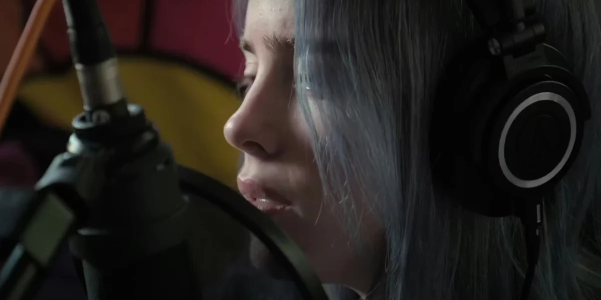 Close-up of Billie Eilish singing softly into a microphone while wearing headphones in a quiet recording setting