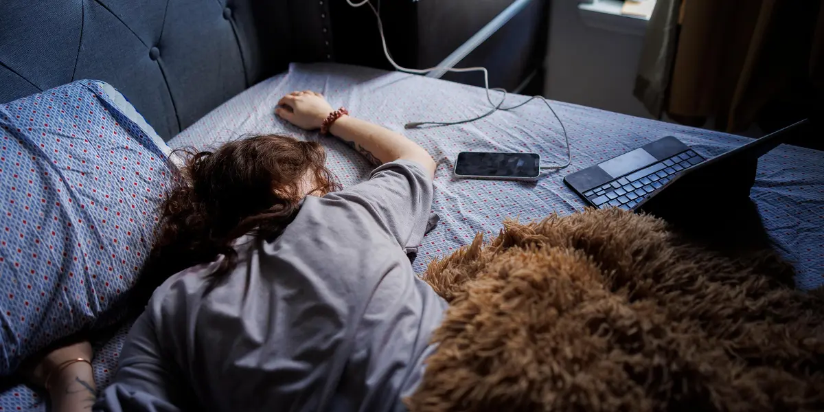 Young person lying on a bed next to a phone and laptop during a late night moment of internet isolation