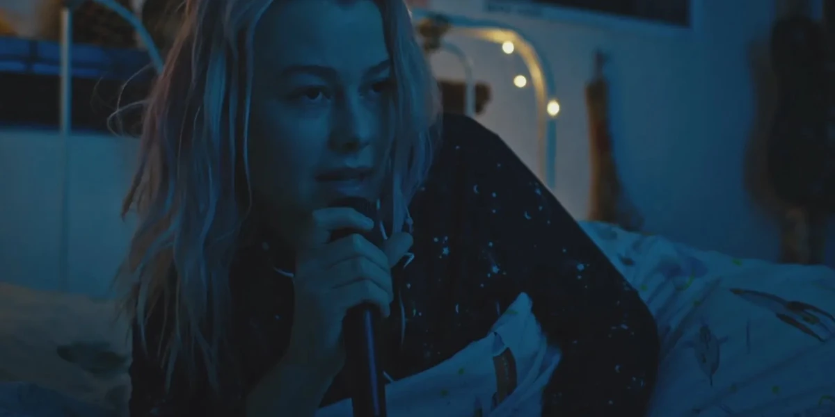 Phoebe Bridgers sitting in a dim blue bedroom atmosphere holding a microphone